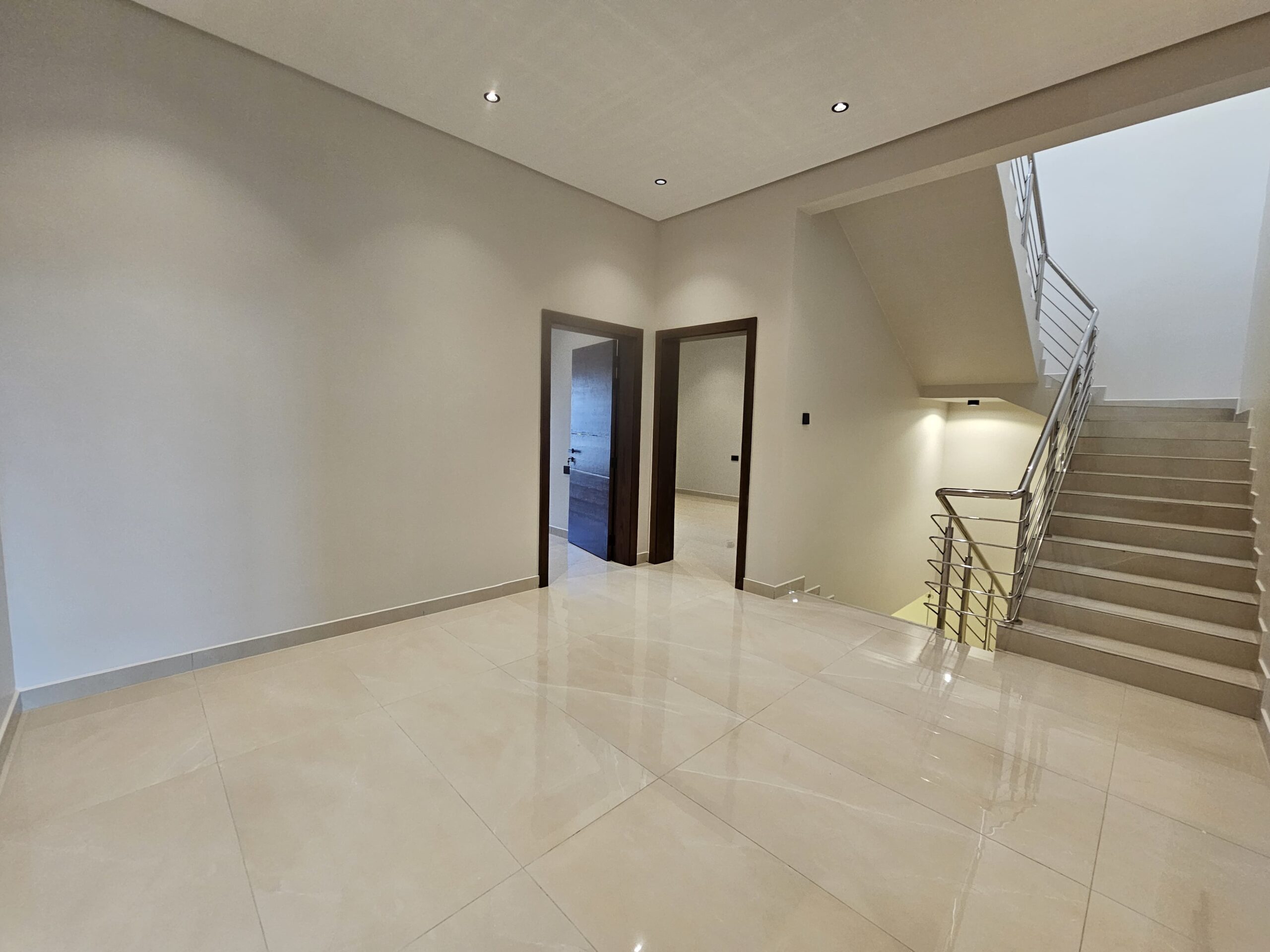 Empty interior with beige tiled floor, two wooden doorways, and a staircase with metal railing. Neutral-colored walls and ceiling with recessed lighting.