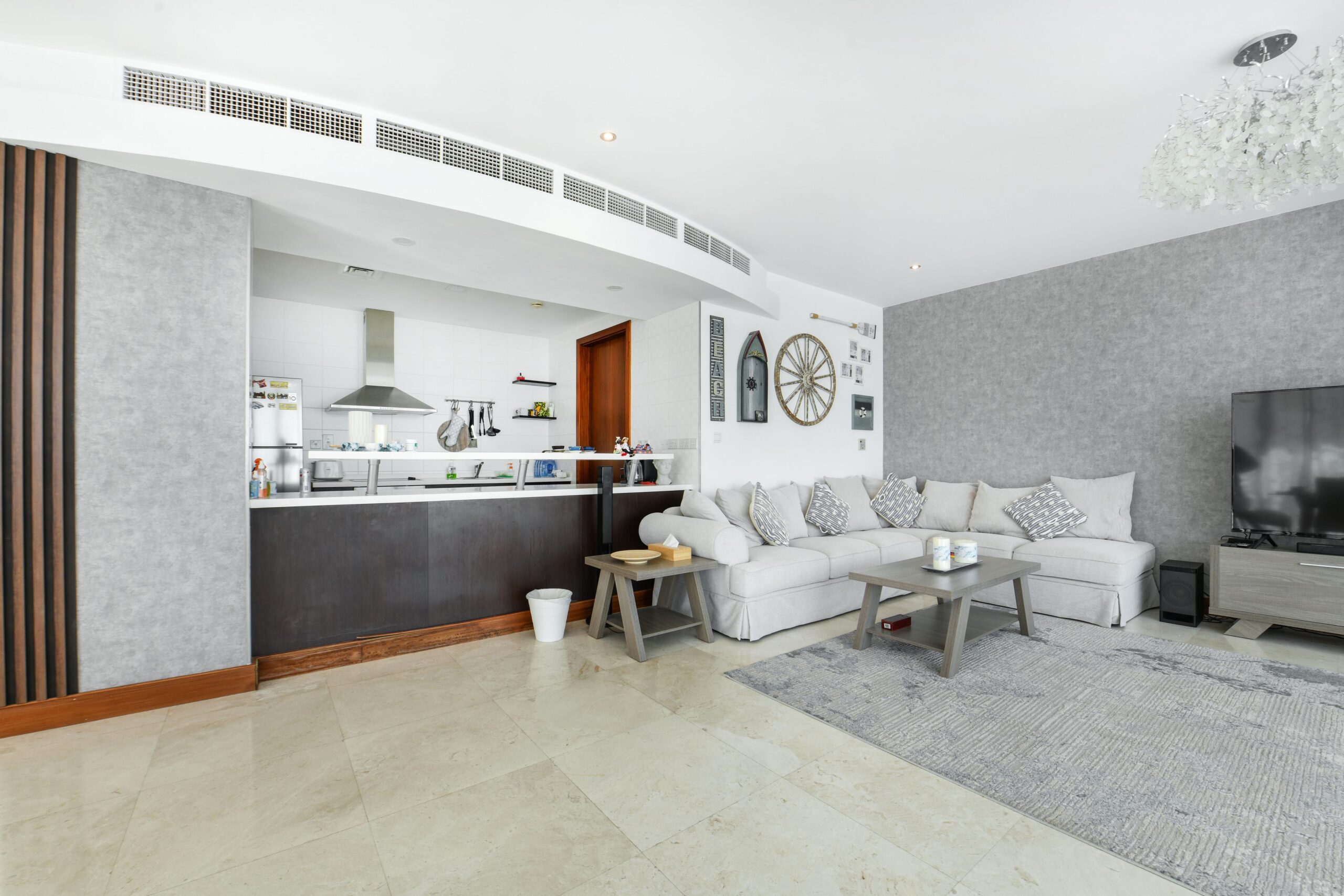 Modern open-plan living room and kitchen with white sectional sofa, gray rug, large TV, kitchen counter with stools, wall decor, and tiled floor.