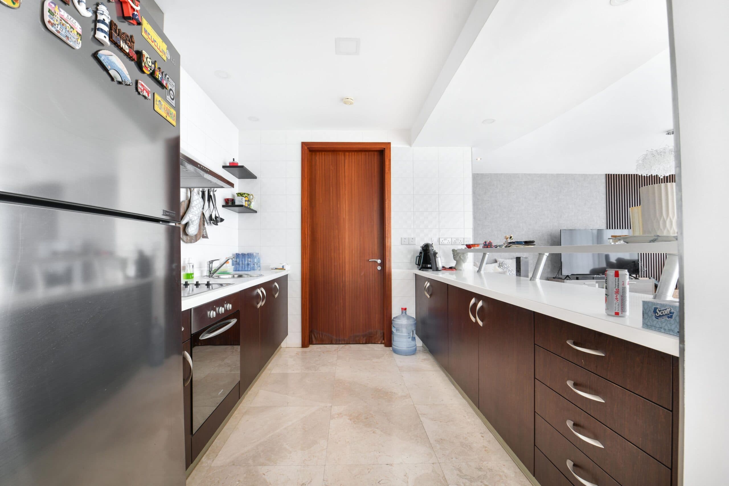 Modern kitchen with stainless steel fridge, dark wood cabinets, white countertops, and beige tile floor. A wooden door at the end, with a wall mirror reflecting the opposite side.