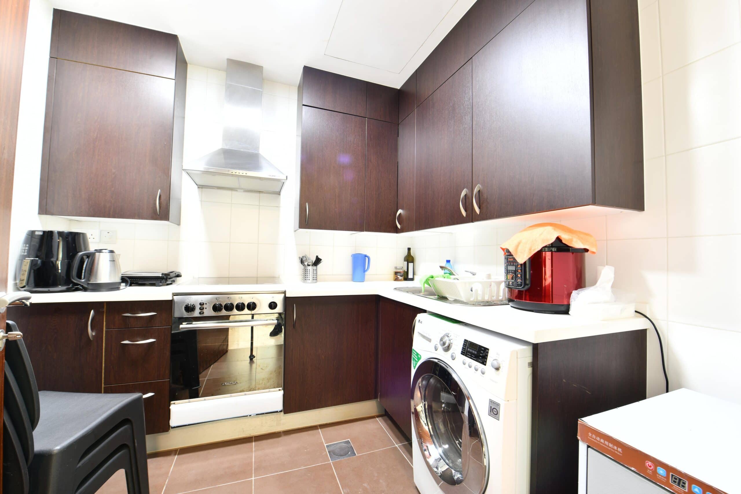 Compact kitchen with dark wooden cabinets, a stainless steel oven, washing machine, and small appliances on the counter. White tiled backsplash and brown floor tiles.