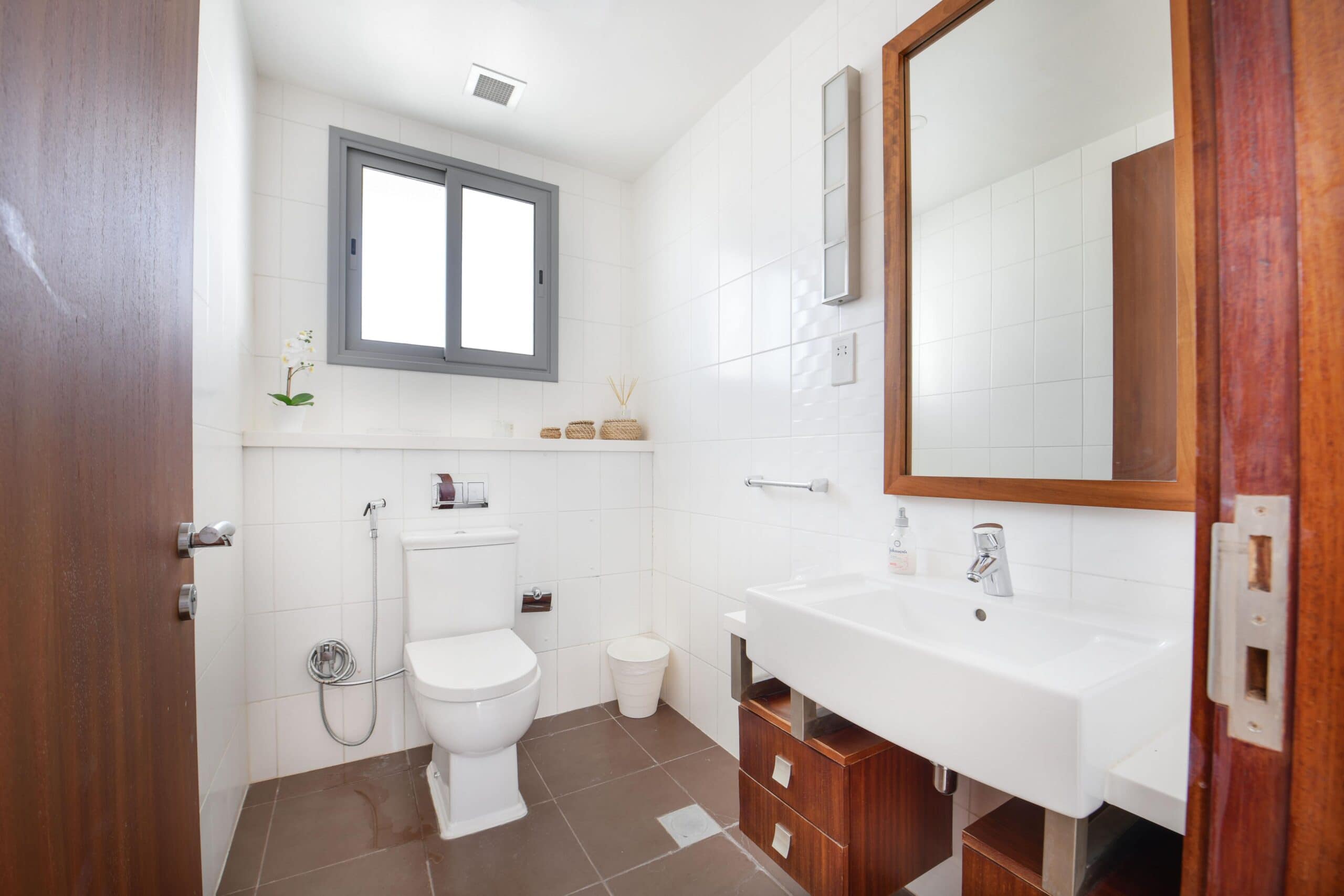 Modern bathroom with a white toilet, white sink, and wooden cabinets. Brown tiled floor, white walls, large mirror, and a window above the toilet. Accessories on shelf and sink.