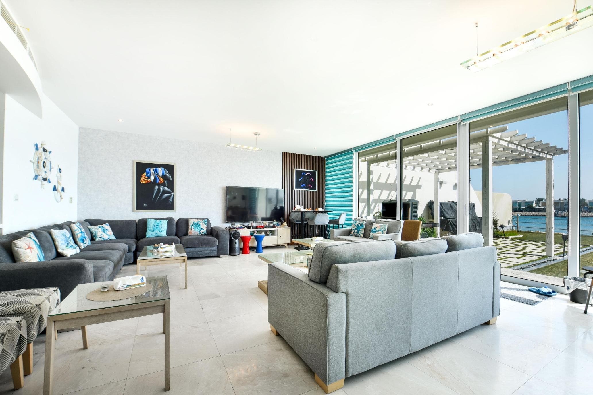 Spacious, modern living room with gray sofas, a large TV, and glass doors leading to a patio overlooking the water. Bright with natural light and contemporary decor.