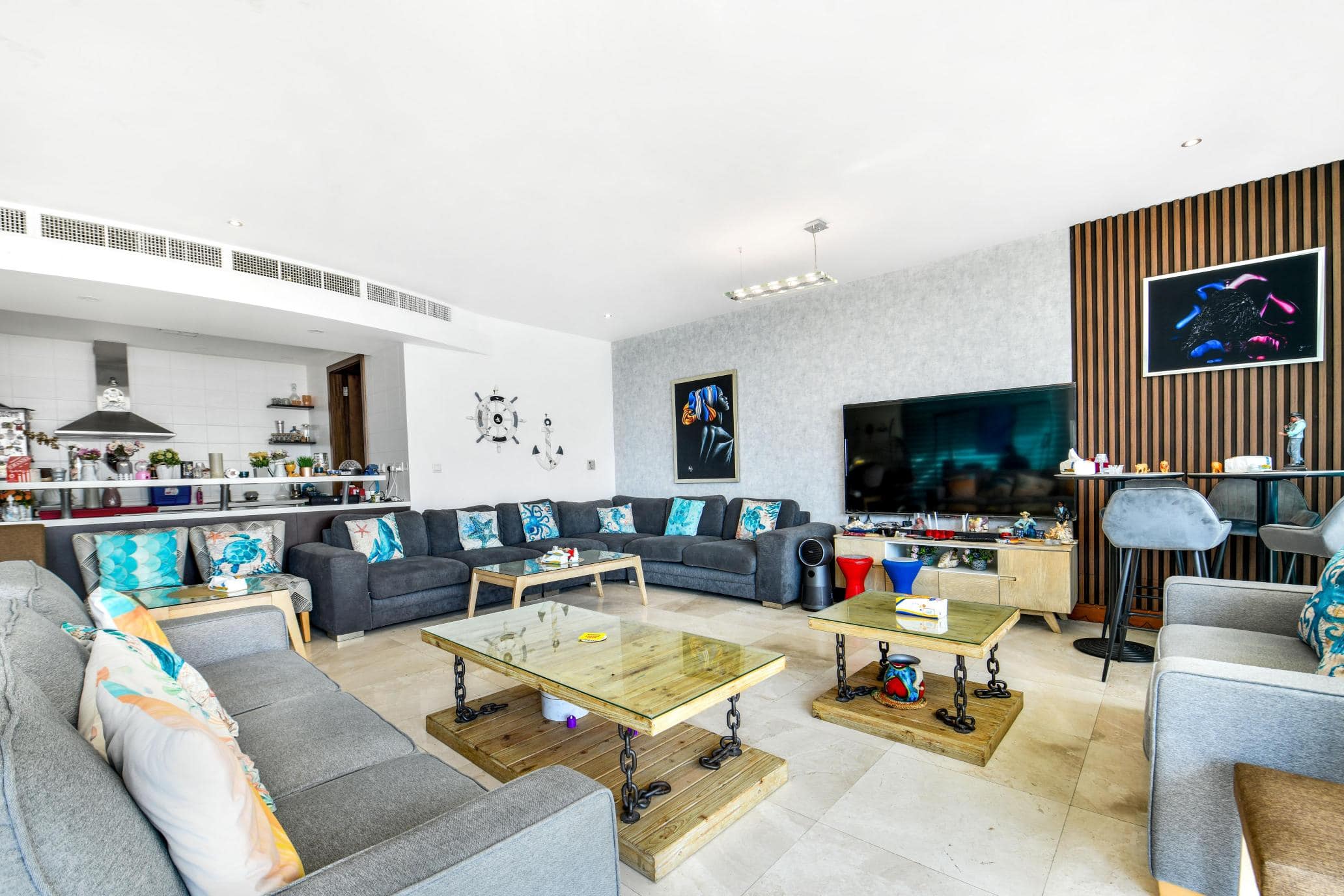 Spacious living room with multiple gray sofas, a large TV, and a glass coffee table. Art decorates the walls. An open kitchen is visible in the background.