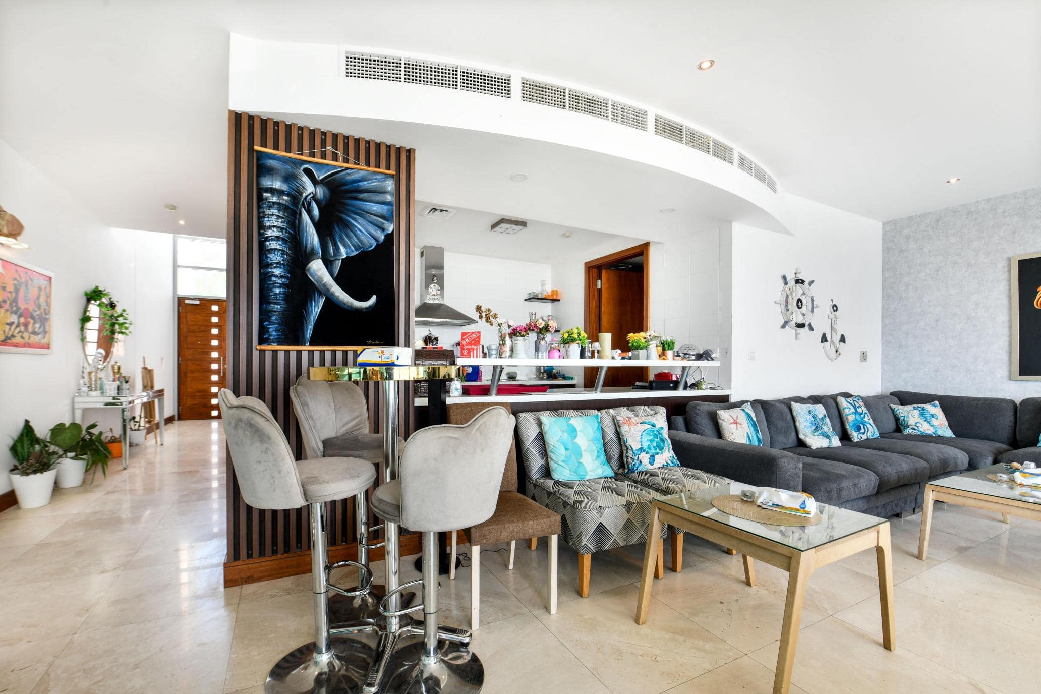 Modern living room with a gray sectional sofa, patterned cushions, and an elephant painting. A kitchen area with bar stools is visible, alongside indoor plants and a light marble floor.
.