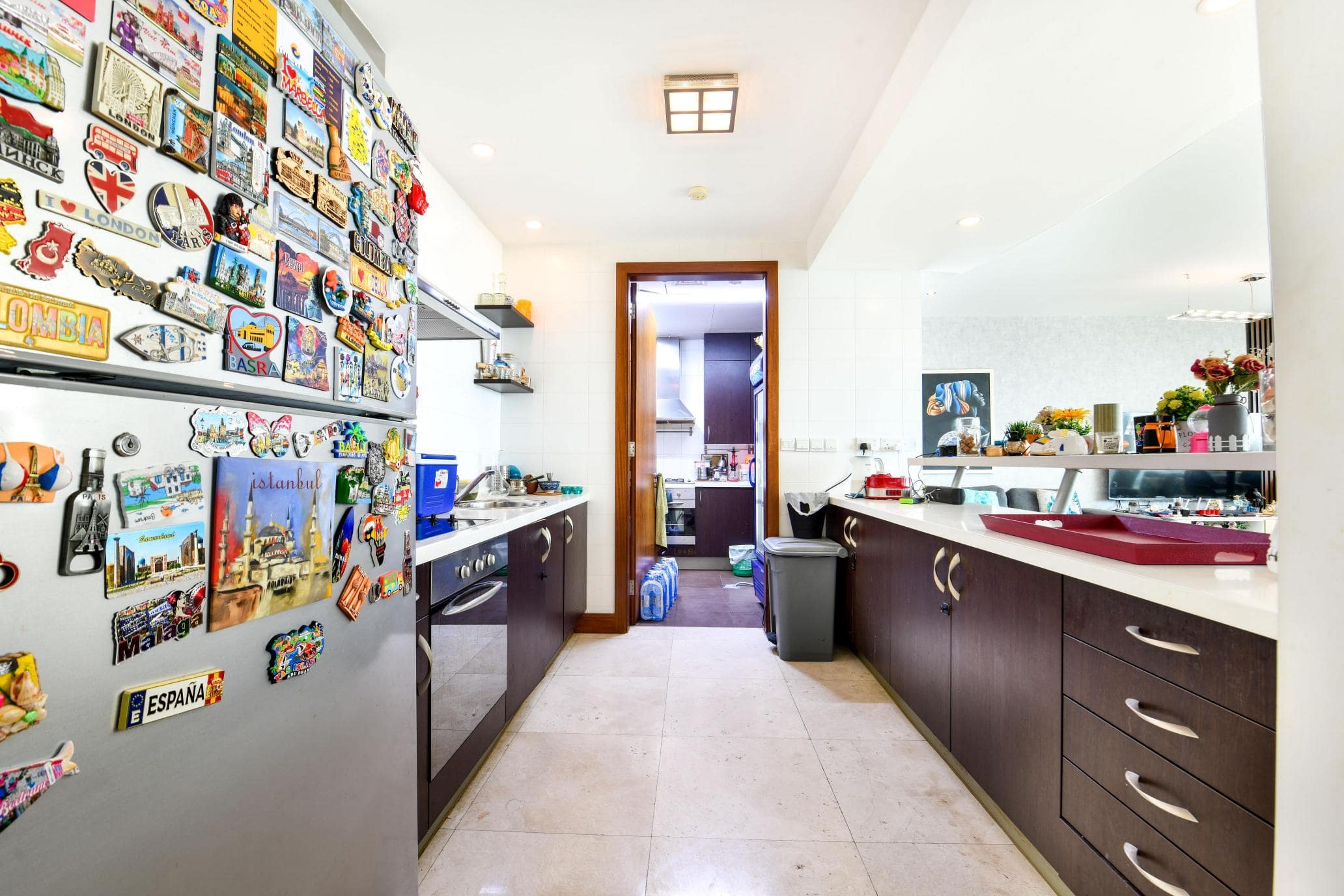 Modern kitchen with dark wood cabinets, stainless steel fridge covered in magnets, light countertops, and a tiled floor. A doorway leads to another room at the back.