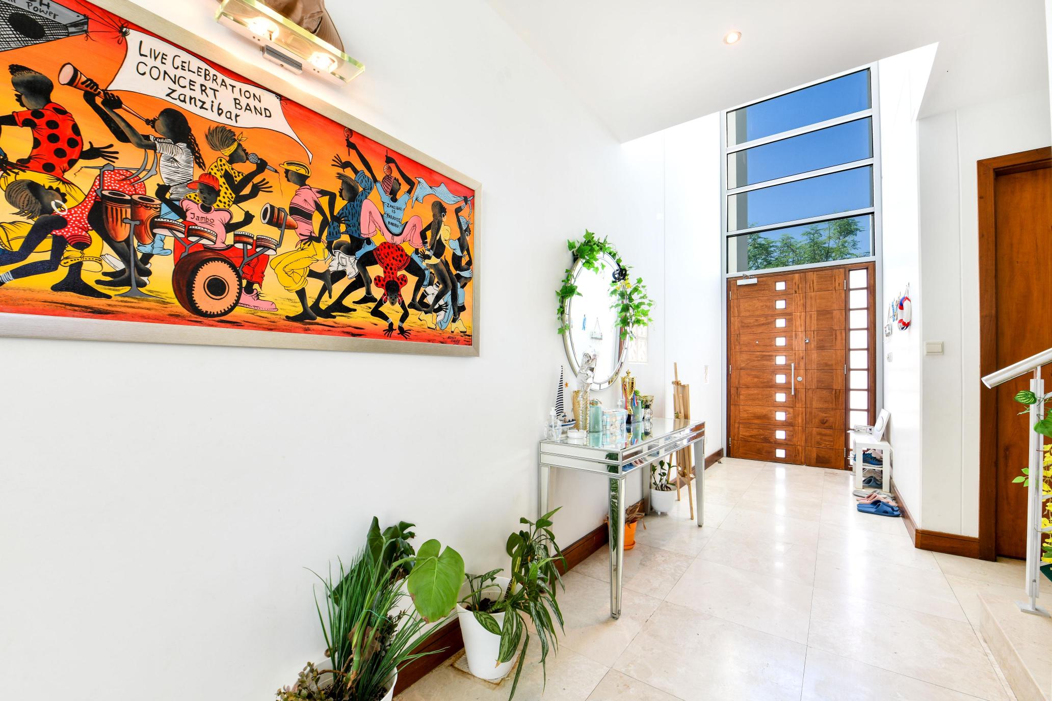 Bright hallway with large colorful painting on the left wall, a console table with a round mirror, and several potted plants. Modern wooden door with horizontal glass panels in the background.