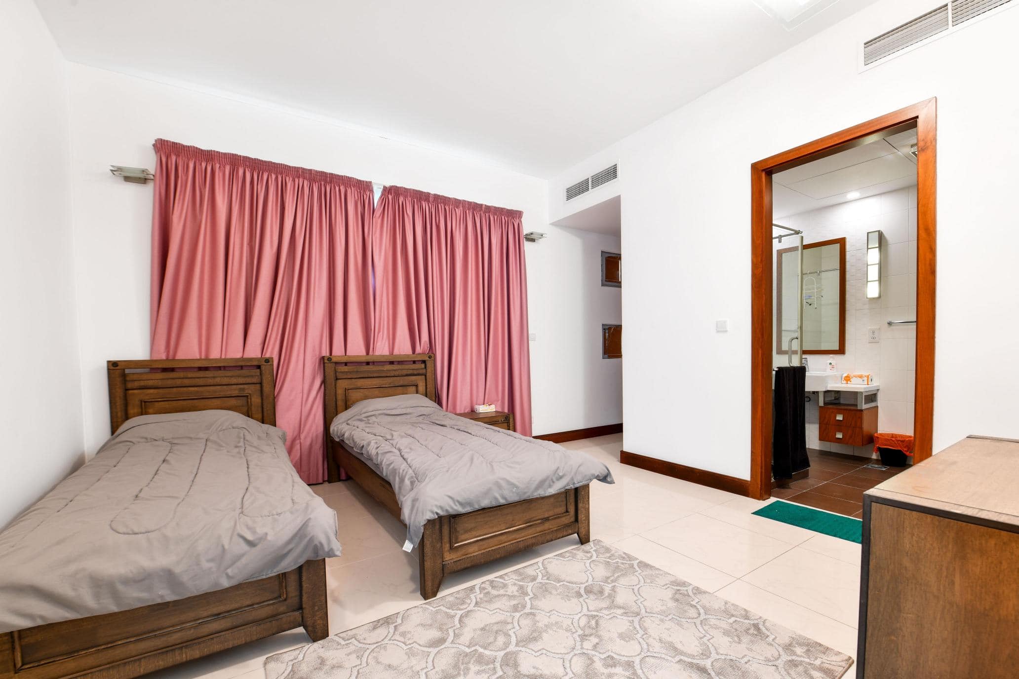 A bedroom with two wooden single beds, gray bedding, pink curtains, a dresser, and an open door leading to a bathroom with a sink and shower.