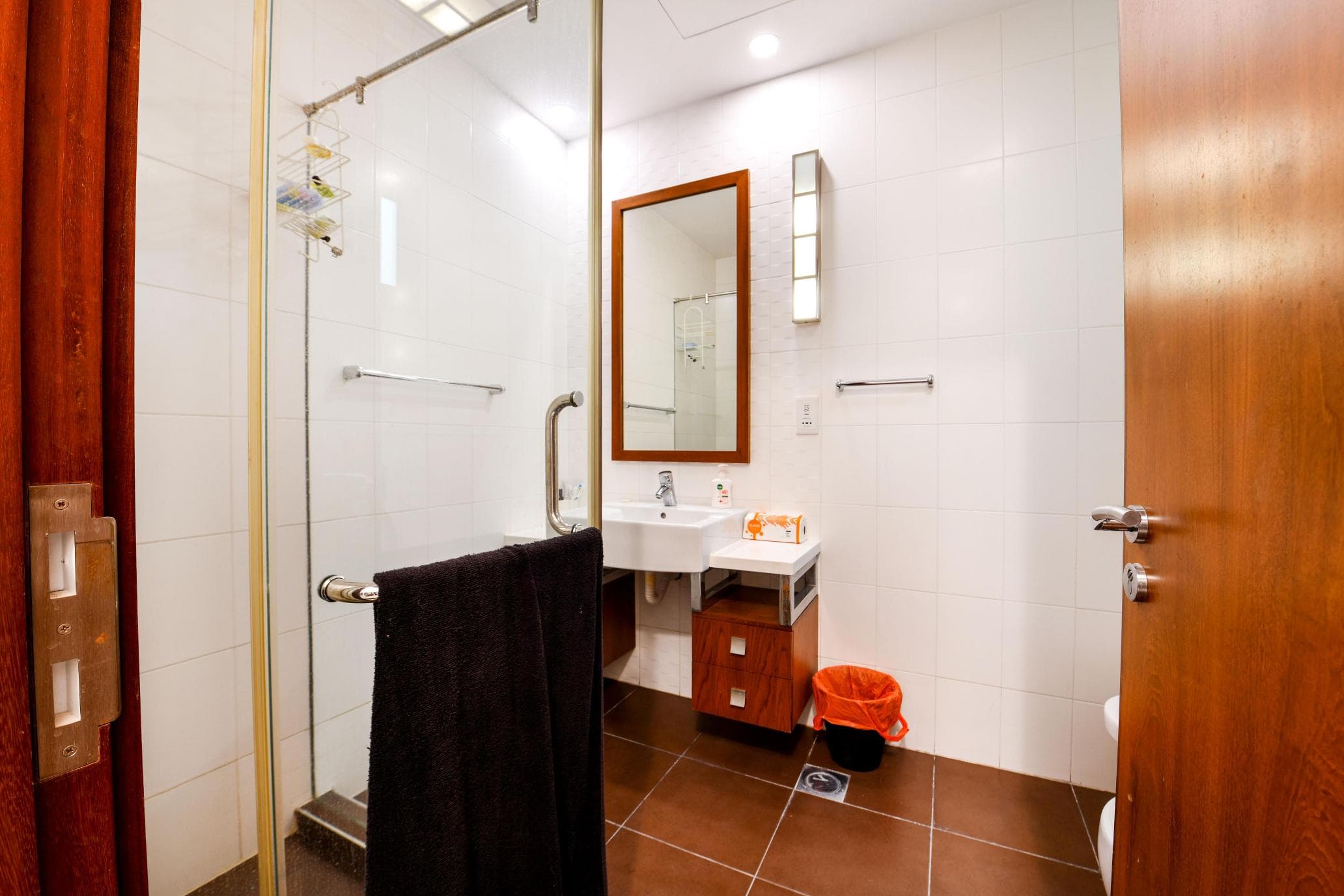 Modern bathroom with glass shower, white tiles, wood accents, a sink with a mirror, towel rack, black towel, and orange trash bin on a brown tiled floor.