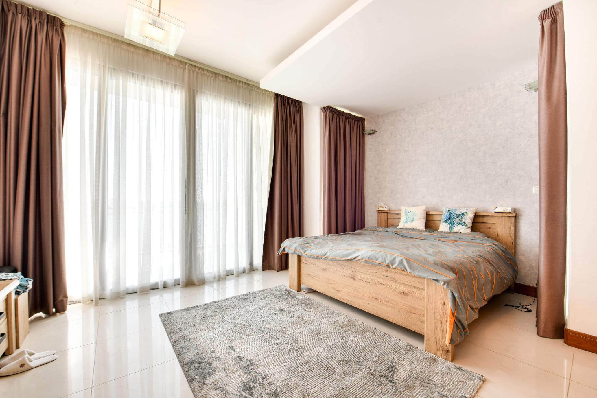 A bedroom with a large wooden bed, gray and blue bedding, windows with sheer and brown curtains, and a gray rug on a glossy floor.