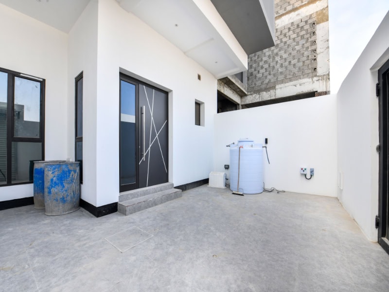 A white outdoor area with a modern glass door, a large water tank, and a blue industrial barrel on a concrete surface.