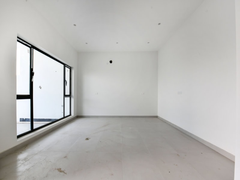 Empty room with white walls, large window on the left, and recessed ceiling lights. The floor is light-colored and slightly dusty.