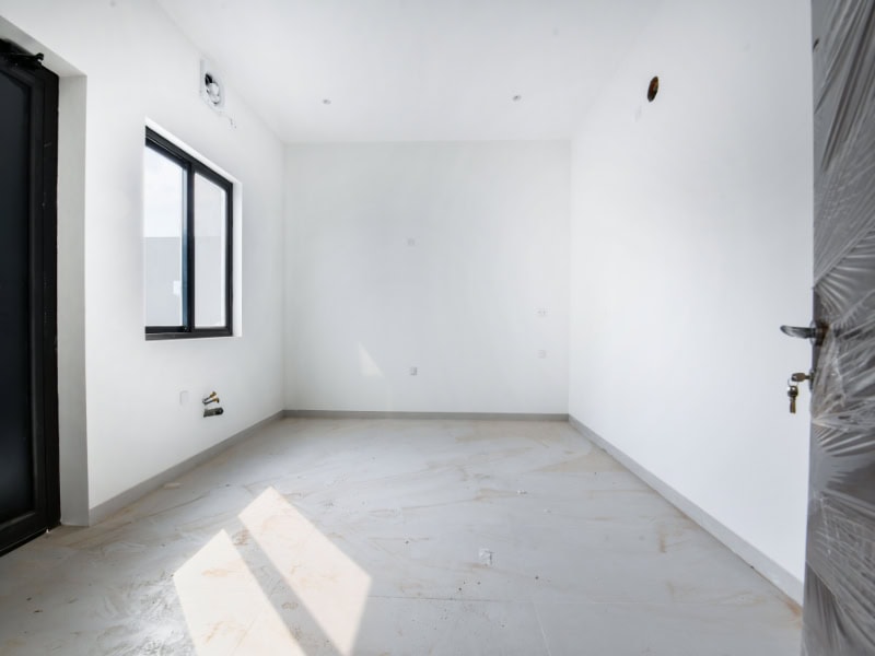 Empty, newly constructed white room with a tiled floor, a window on the left wall, and a black door on the right side. Dust and plaster remnants are scattered on the floor.