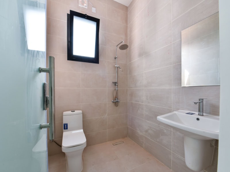 Minimalist bathroom with beige tiles, featuring a toilet, wall-mounted shower, rectangular mirror, and a sink with a silver faucet. A frosted glass door and a small window provide light.