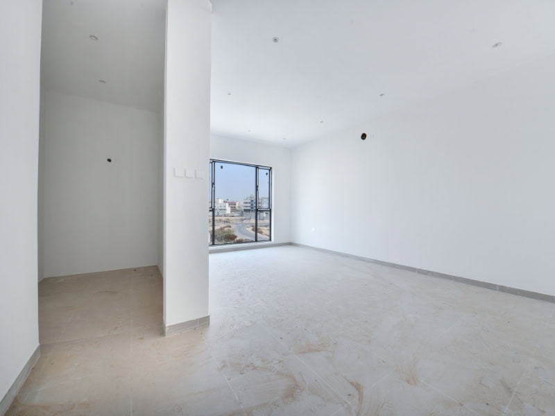 Empty white room with a large window, beige tiled floor, and recessed ceiling lights. An adjacent small area to the left has bare walls and an uninstalled fixture.