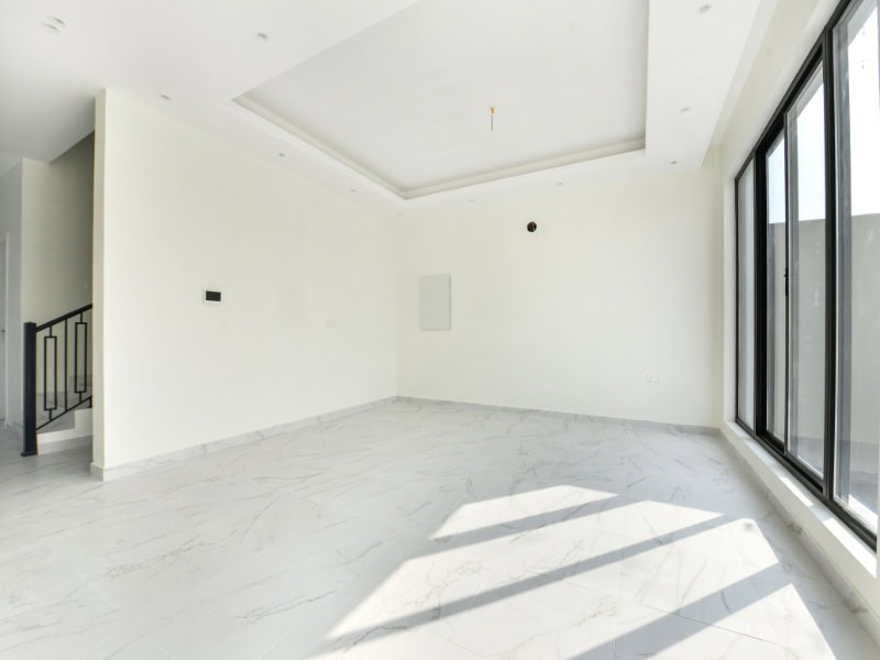 An empty room with white walls, marble floor, large windows on the right, and a partially visible staircase to the left.