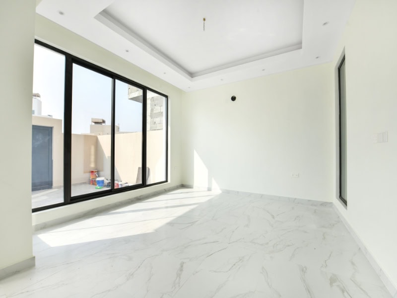 Empty room with large windows, white walls, and marble floor. Sunlight streams in, casting shadows on the floor. Boxes and toys visible outside the window on a balcony.