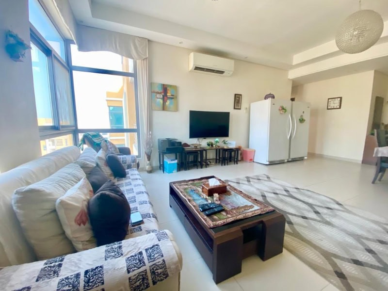 Bright living room with a couch, coffee table, TV, and wall decor. Large window, air conditioner, and refrigerator visible. Neutral colors and patterned textiles decorate the space.