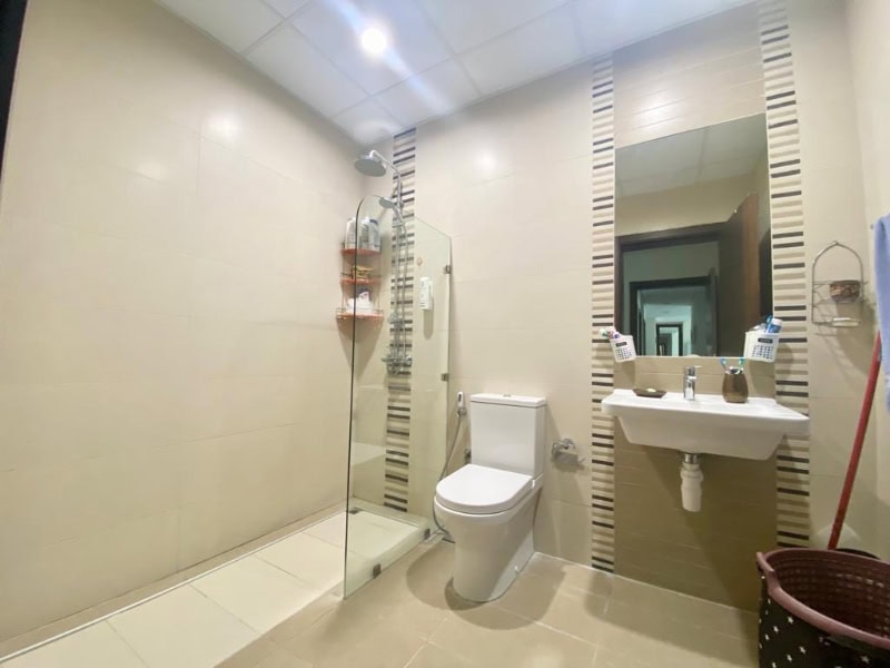 Modern bathroom with a glass-enclosed shower, toilet, and wall-mounted sink. Tiled walls and floor, shelves with toiletries, and a wastebasket complete the scene.