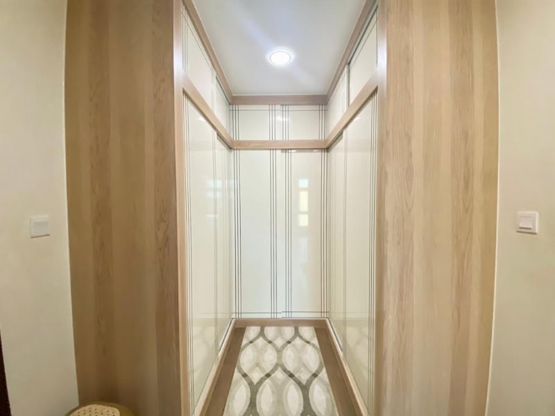 A narrow, well-lit hallway with light wood paneling and patterned floor, leading to a slightly reflective door at the end.