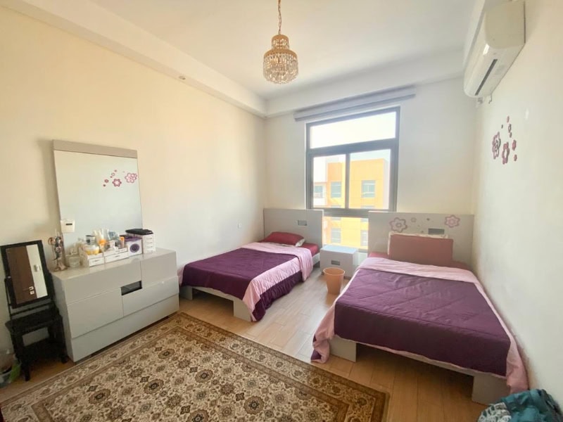 A tidy bedroom with two single beds, purple bedspreads, a dresser with a mirror, and a window letting in natural light. A chandelier hangs from the ceiling.