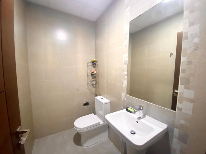 A small bathroom with beige tiles, featuring a toilet, a white sink with a faucet, a large mirror, and a metal rack with toiletries on the wall.