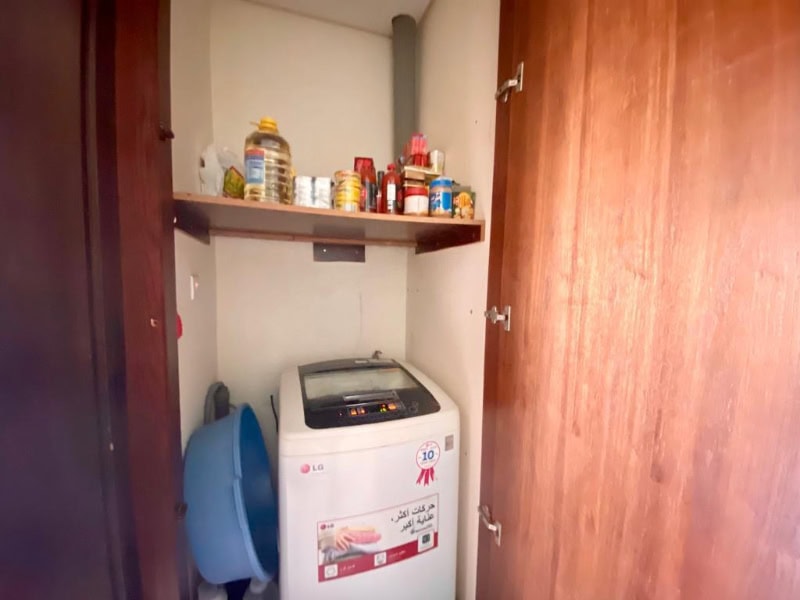 Small laundry closet with a top-loading washing machine, blue laundry basket, and a shelf above holding various canned goods and bottles. Wooden doors frame the closet.