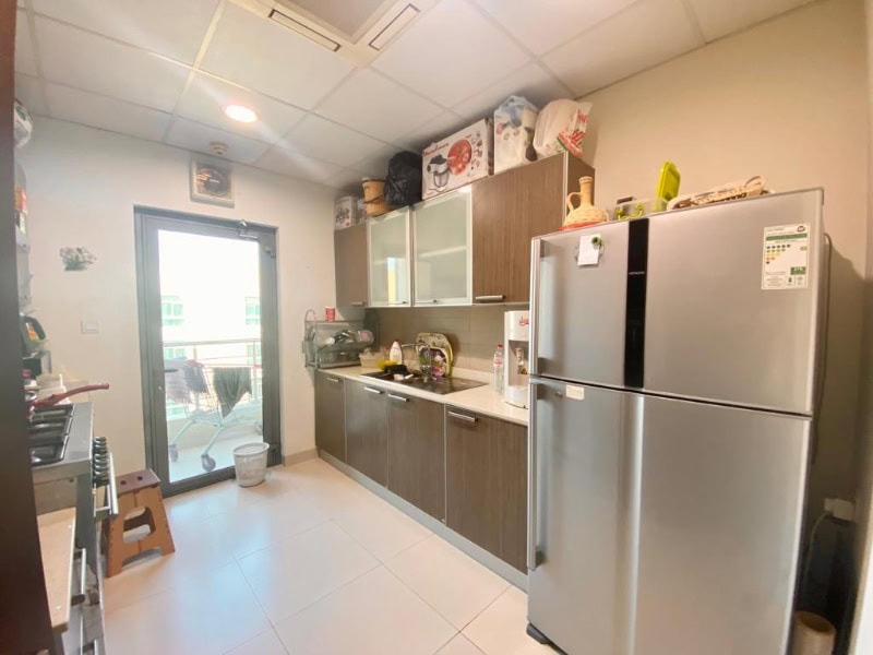 Modern kitchen with wooden cabinets, stainless steel fridge, countertop appliances, and a washing machine. A glass door leads to a balcony with visible buildings outside.