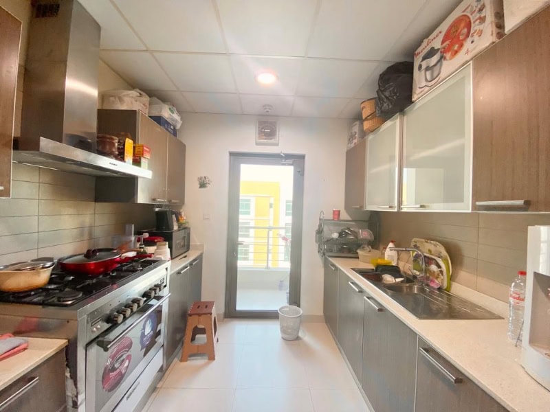 A modern kitchen with wooden cabinets, a gas stove with cooking utensils, a sink on the right, and a glass door leading to a small balcony. Various kitchen items are visible on the counters.