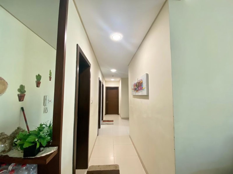 A narrow hallway with cream-colored walls and a tiled floor. There are three dark wooden doors and a wall-mounted artwork. Potted plants are on a small shelf to the left.