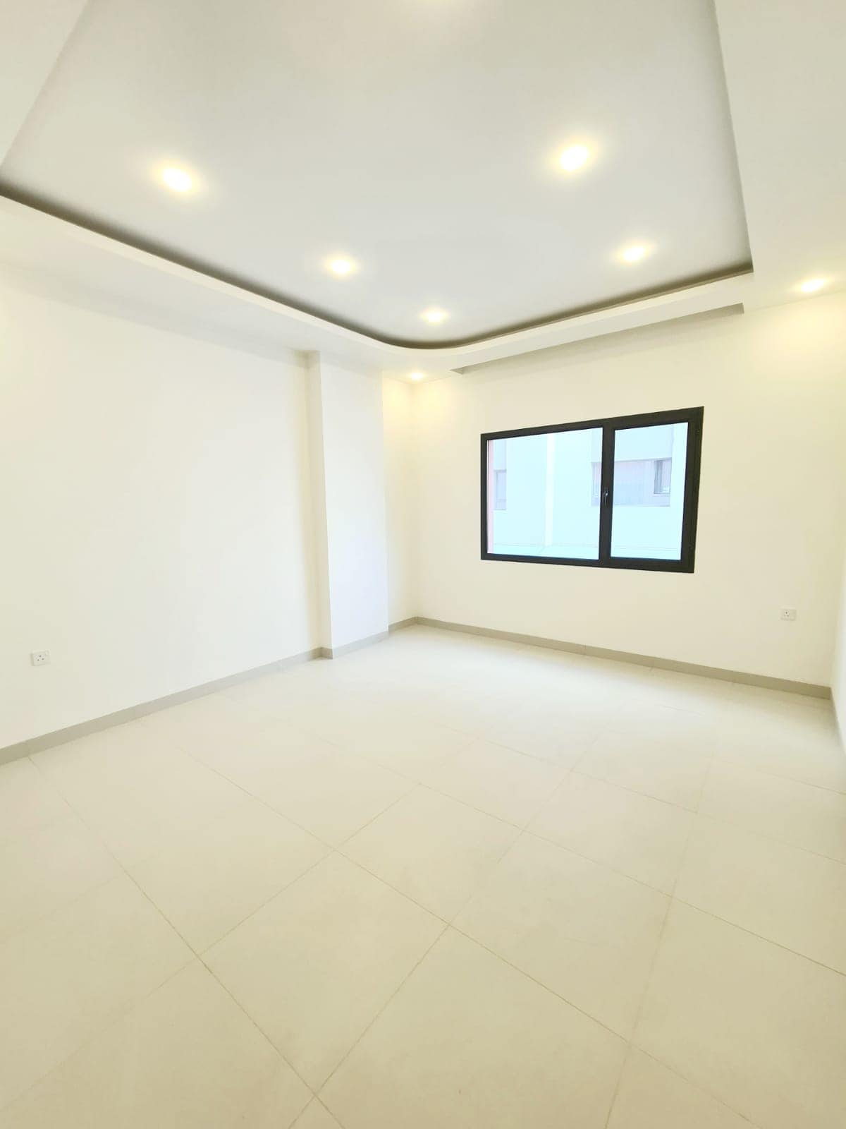 Empty room with light-colored tiled floor, white walls, recessed ceiling lights, and a window with a black frame on one wall.