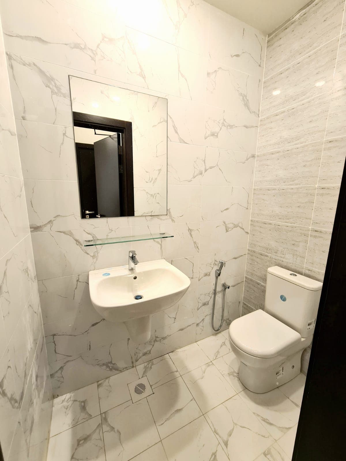 White bathroom with a marble-patterned tile design, featuring a wall-mounted sink, a mirror above, a toilet, and a handheld bidet spray on the side.