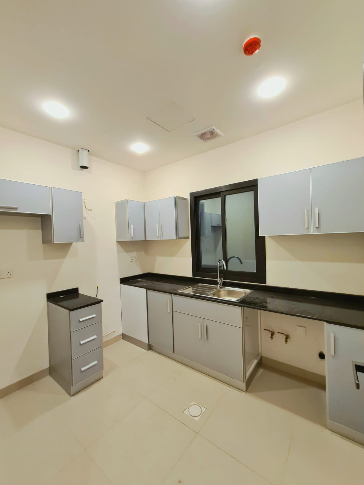 A modern kitchen with light gray cabinets, a black countertop, and a stainless steel sink. The floor is tiled, and the room is well-lit with recessed ceiling lights.