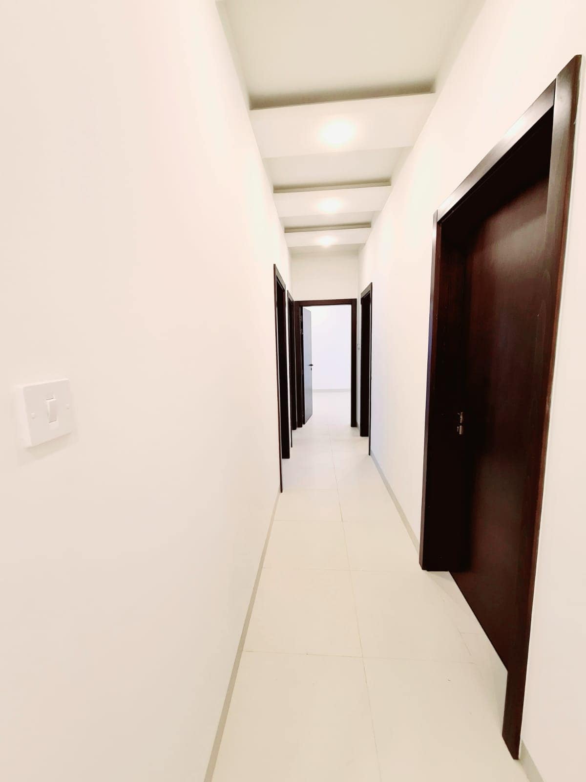 Long narrow hallway with white walls and tiled floor, leading to a series of dark wood doors on both sides. Ceiling features recessed lighting.