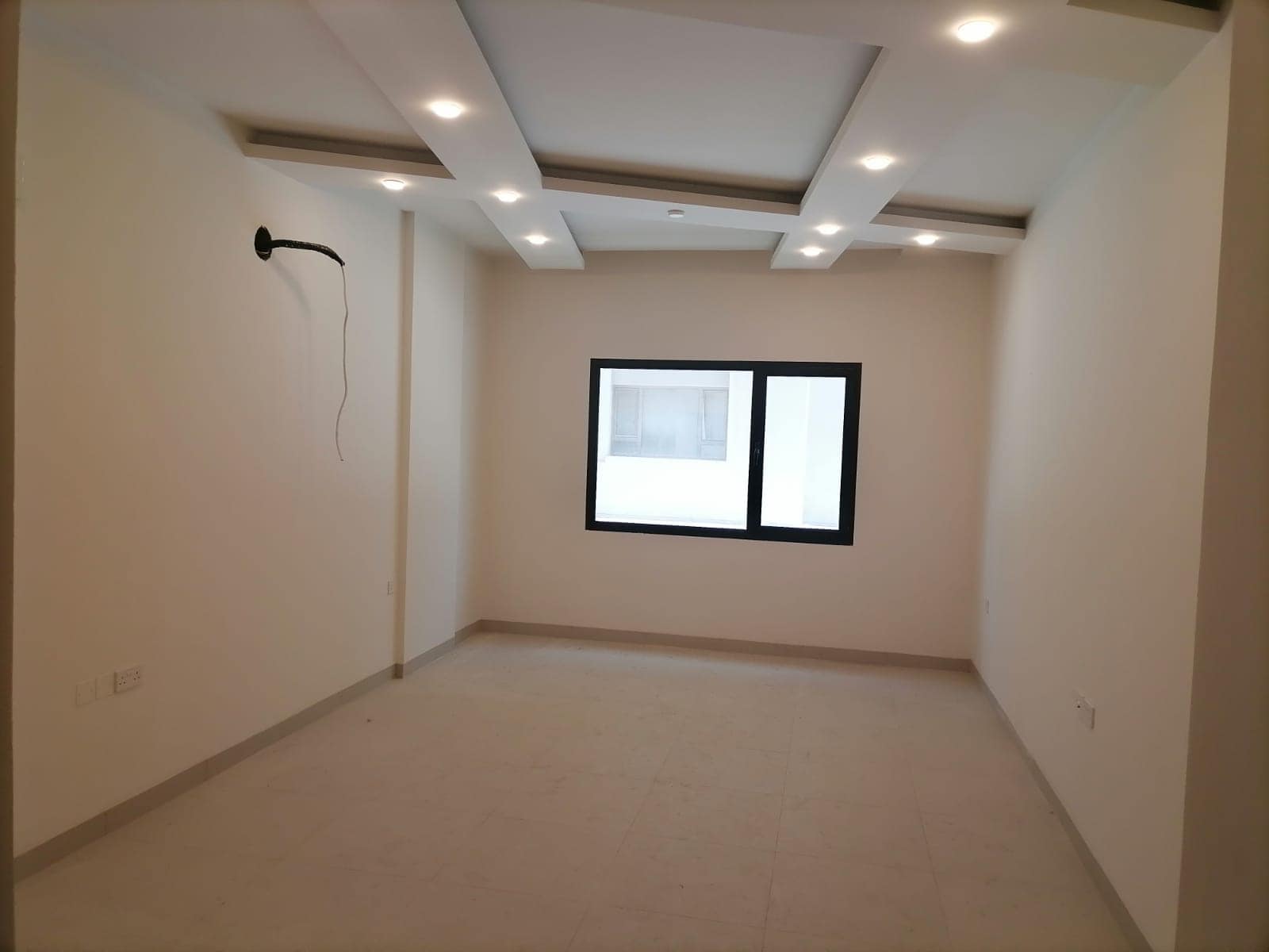 Empty room with a beige tiled floor, white walls, recessed ceiling lights, a wall-mounted TV bracket, and a window showing an exterior building.