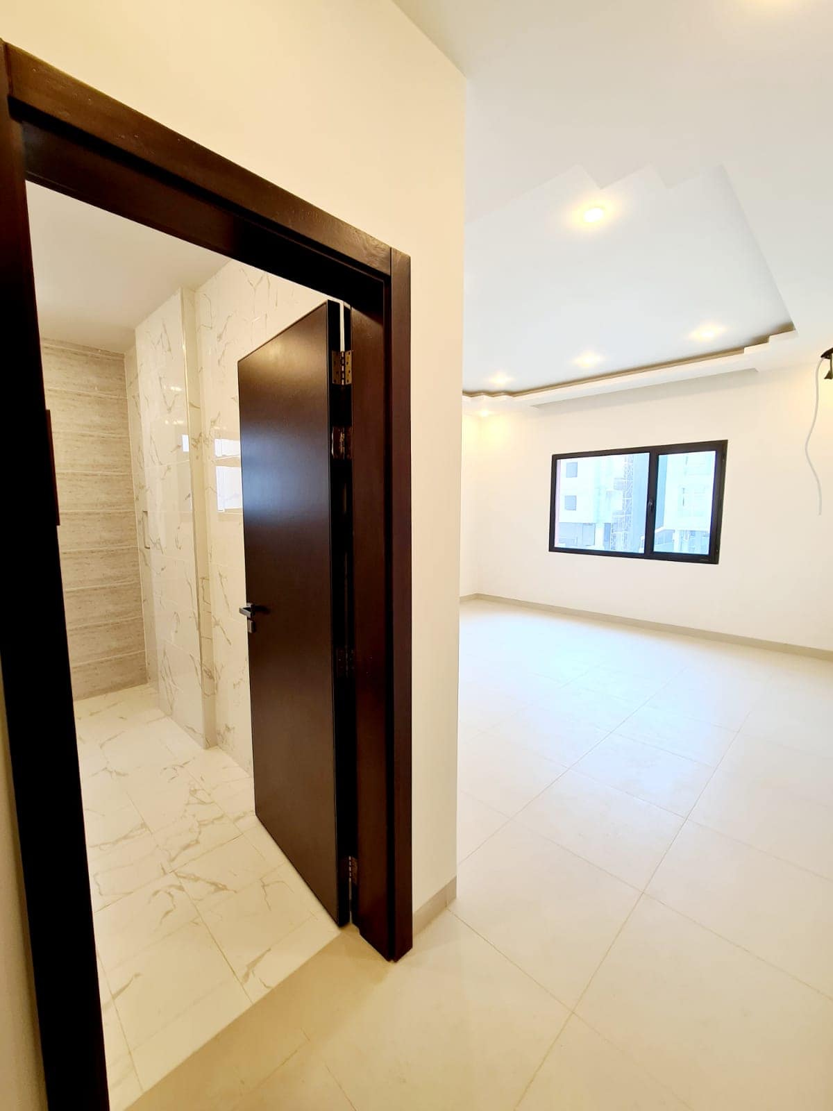 Minimalist interior with a partially open wooden door leading to a tiled room. The adjacent space features white walls, large windows, and recessed ceiling lights.