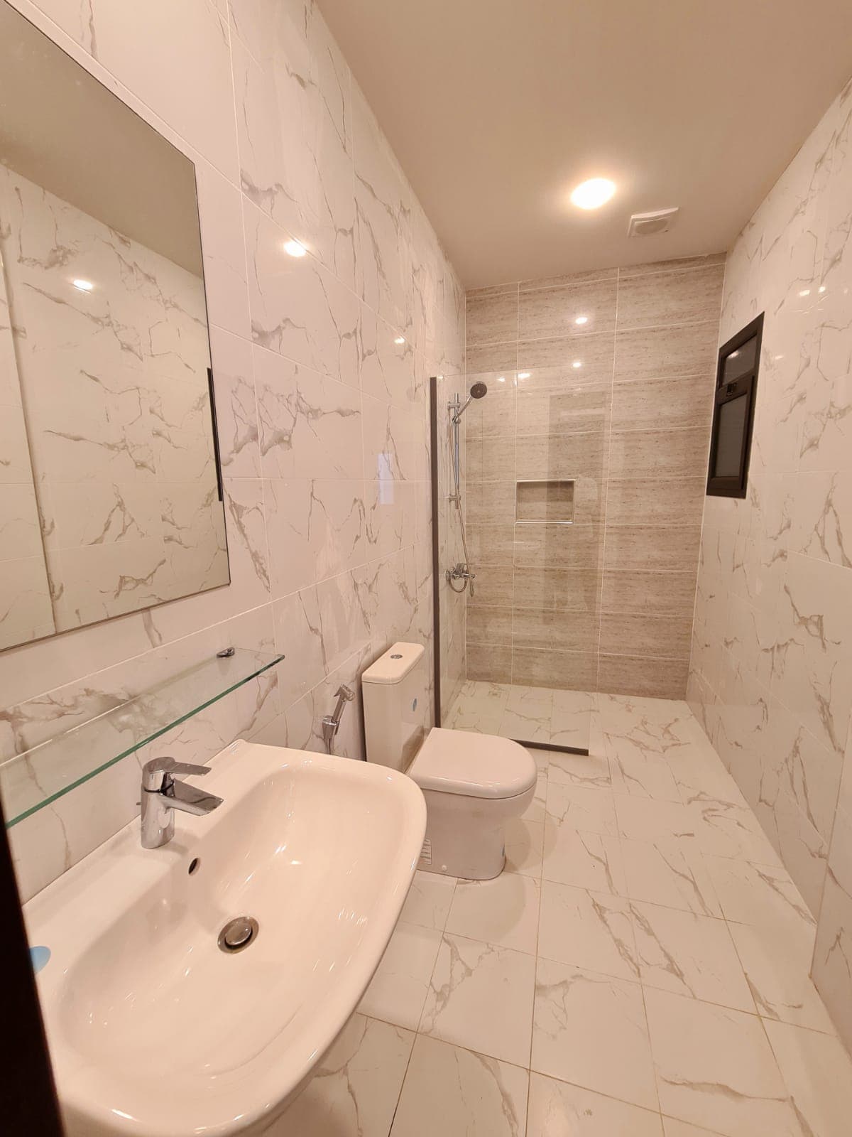 Modern bathroom with marble-patterned tiles, a wall mirror above a white sink, a toilet, and a glass-enclosed shower area.