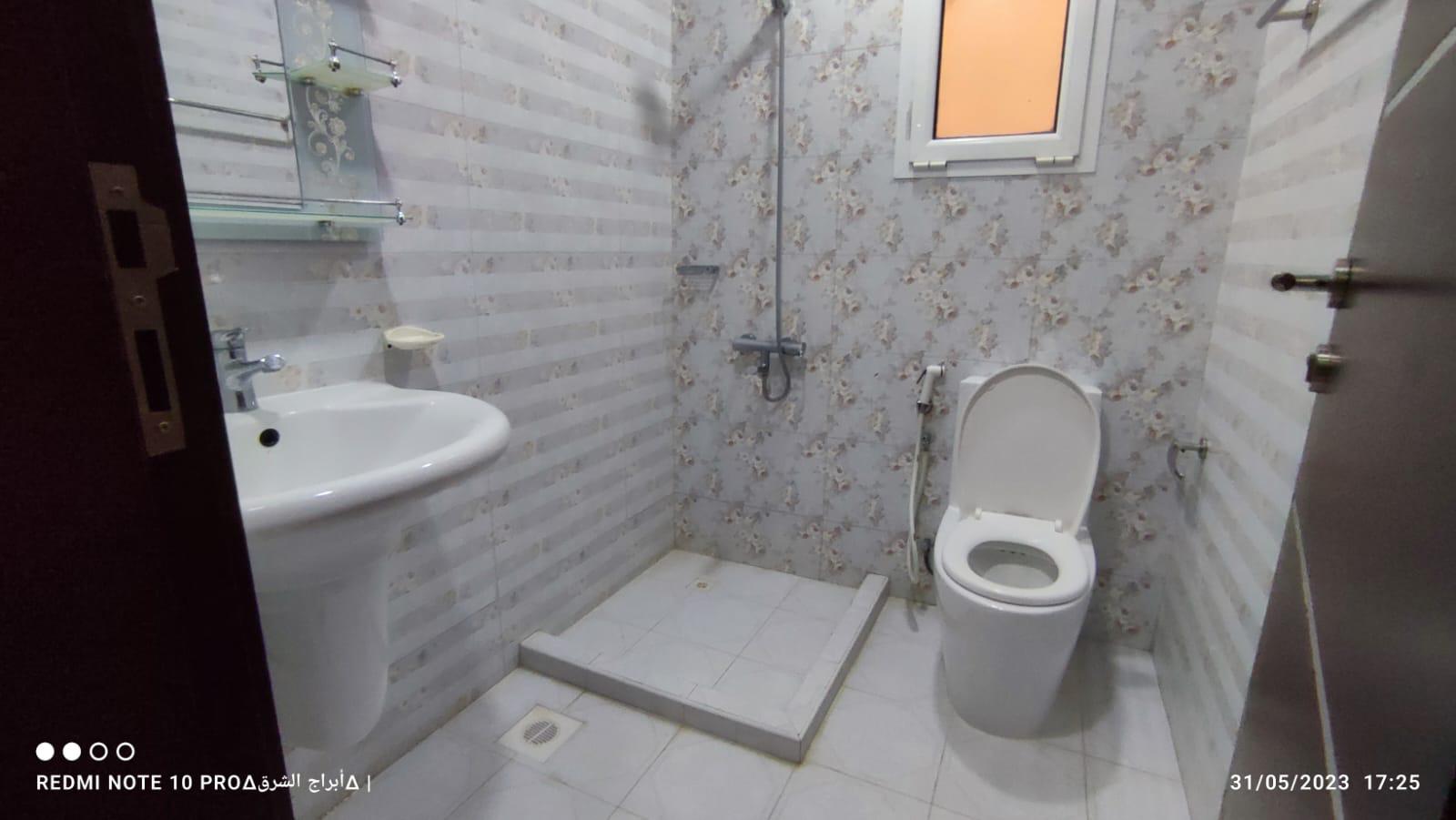 A bathroom with patterned walls, a sink, a toilet, and an open shower area. A small window is above the toilet, and the floor is tiled.