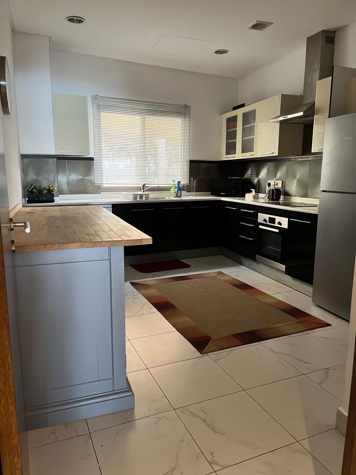 Modern kitchen with black cabinets, stainless steel appliances, wooden countertop, and a beige rug on white tile floor. A window with blinds is above the sink.