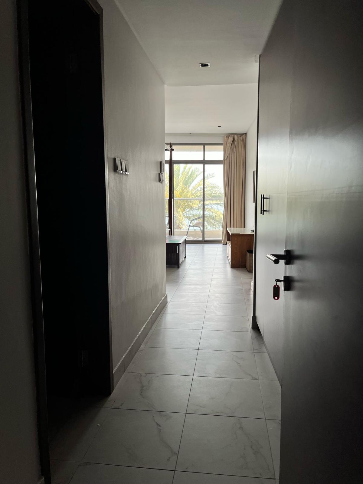 View of a hotel room entrance with tiled floor, leading to a window and balcony. Palm trees are visible outside.