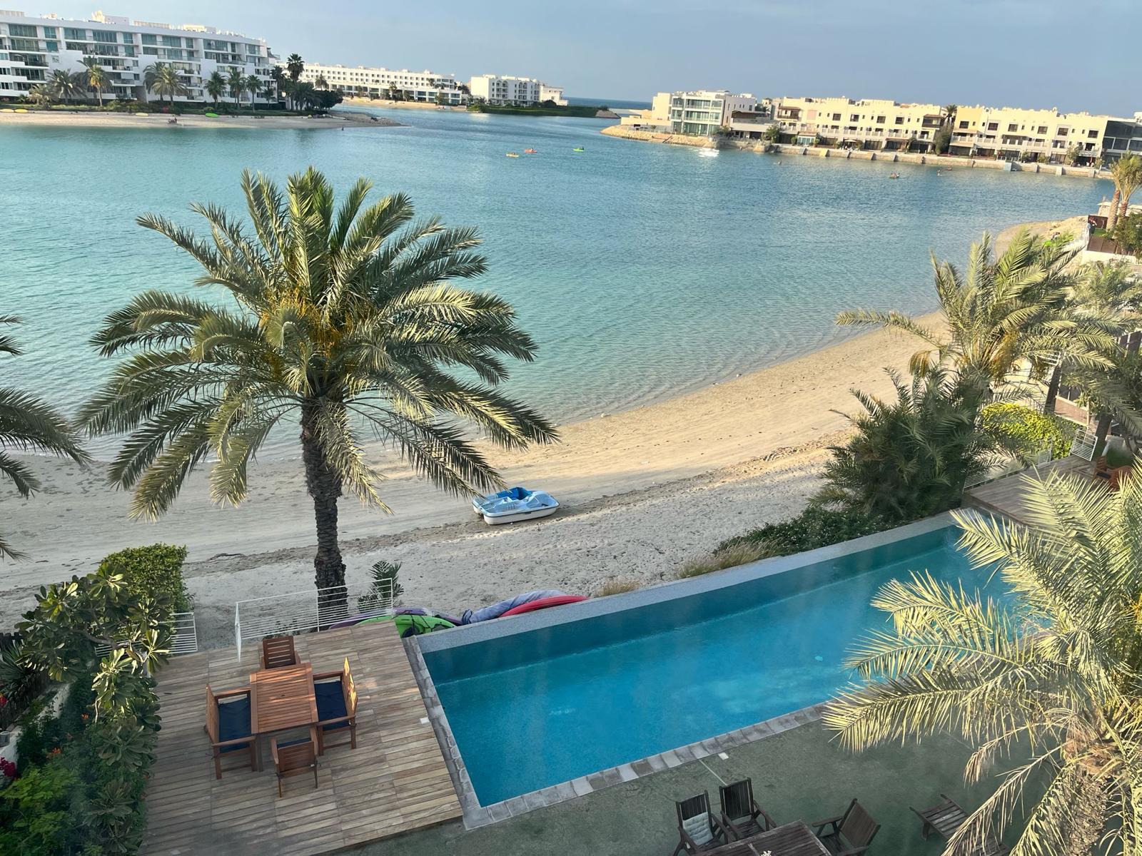 View from above of a beachside area with palm trees, a pool, and a sandy shore. Buildings and a calm body of water are visible in the background.