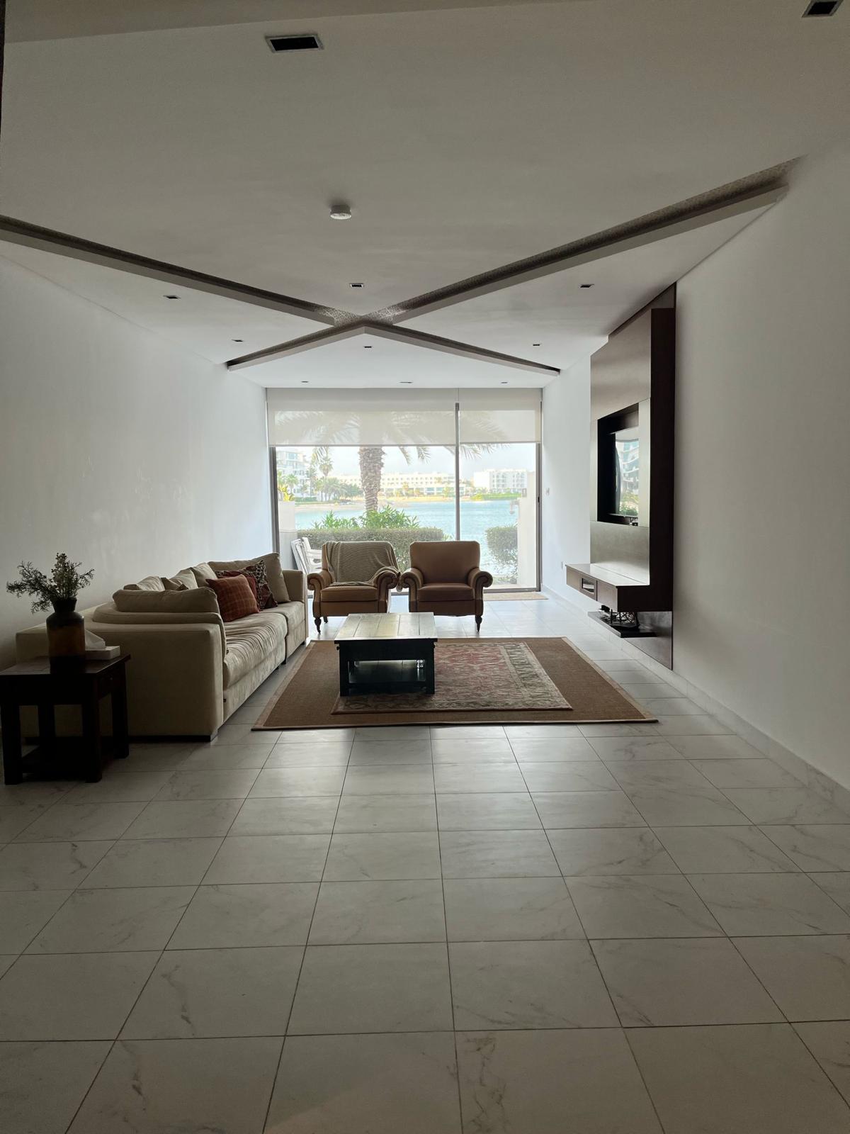 A modern, minimalistic living room with a sofa, armchairs, a coffee table on a rug, and a wall-mounted TV. Large sliding glass doors open to a view of trees and the outdoors.