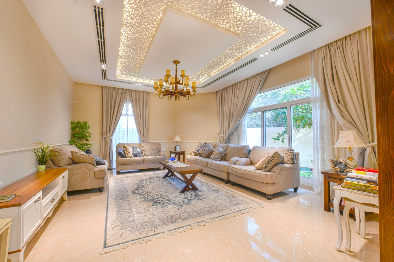 A modern living room with a large rug, two beige sofas, a wooden coffee table, and an ornate ceiling with a chandelier. Large windows with curtains allow natural light.