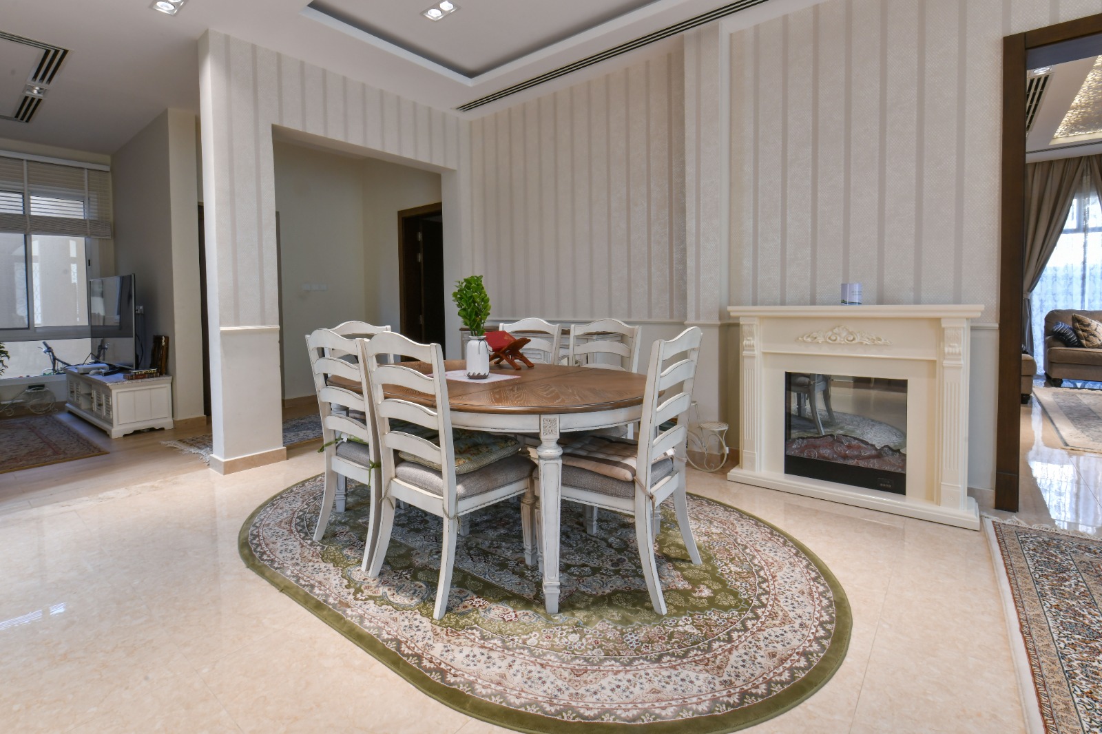 Dining room with a round wooden table, six white chairs, and a decorative rug. A fireplace is on the right, and adjacent rooms are visible through open doorways.