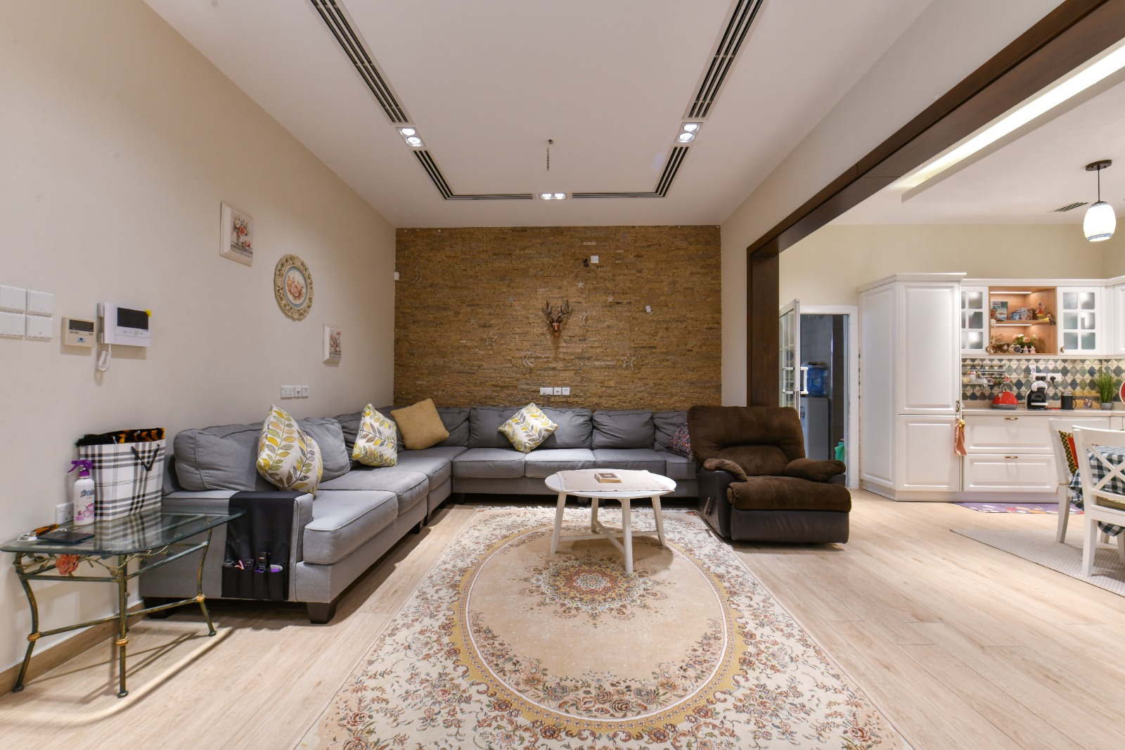 A modern living room with a gray sectional sofa, a round coffee table, and a recliner. There's a decorative rug on the wooden floor and an open view into the kitchen area.