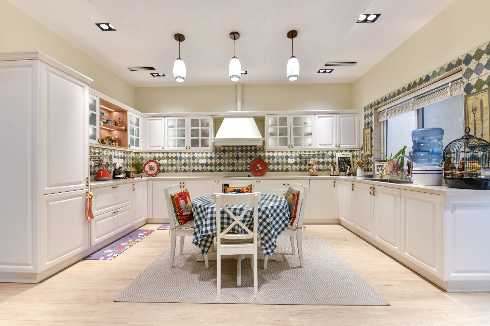 Modern kitchen with white cabinets, island, and dining area. Geometric backsplash, pendant lights, and wood flooring. Water dispenser and birdcage on counter.