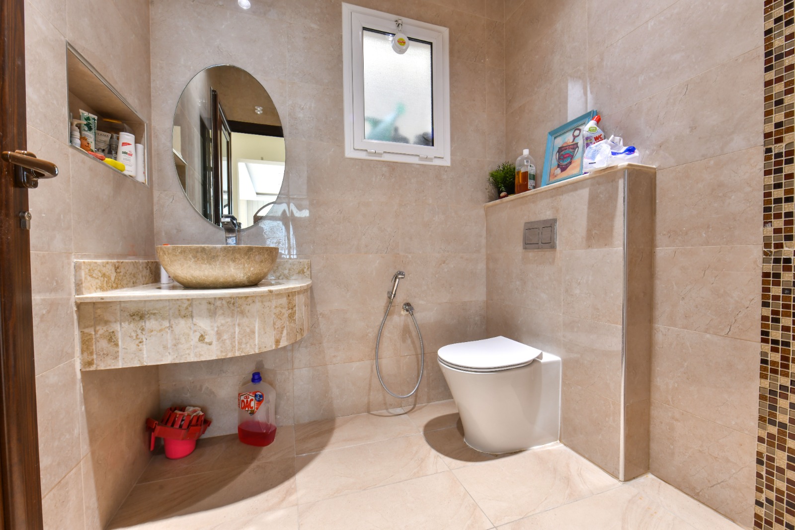 A modern bathroom with beige tiles, featuring a circular mirror, a bowl sink, and a wall-mounted toilet. A small shelf holds toiletries, and a bidet spray is next to the toilet.