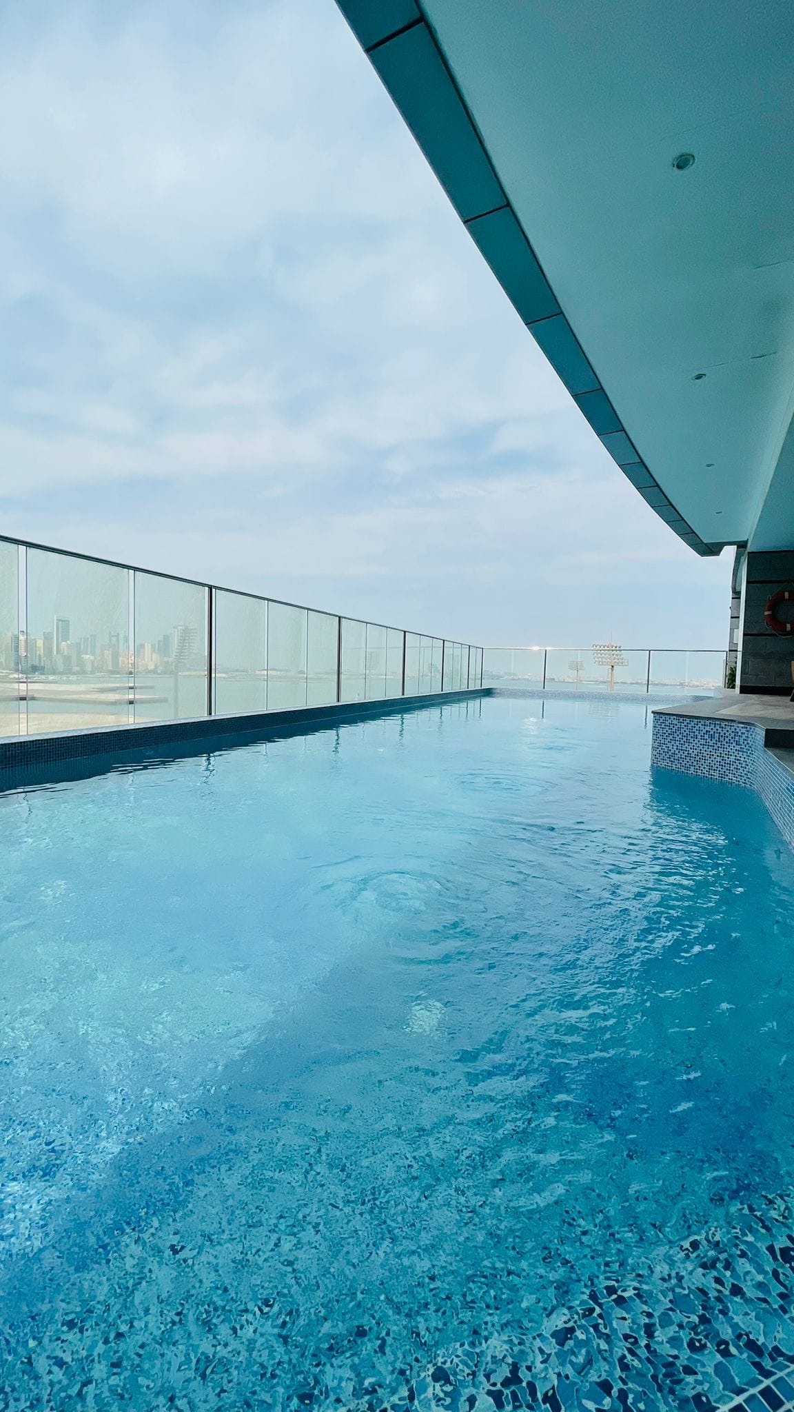 Rooftop infinity pool with clear blue water, glass railing, and cityscape view under a partly cloudy sky.