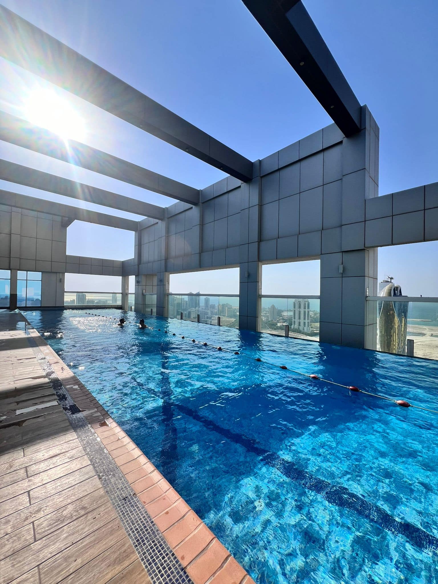 Rooftop swimming pool under a clear blue sky, with city views in the background and sunlight shining brightly.