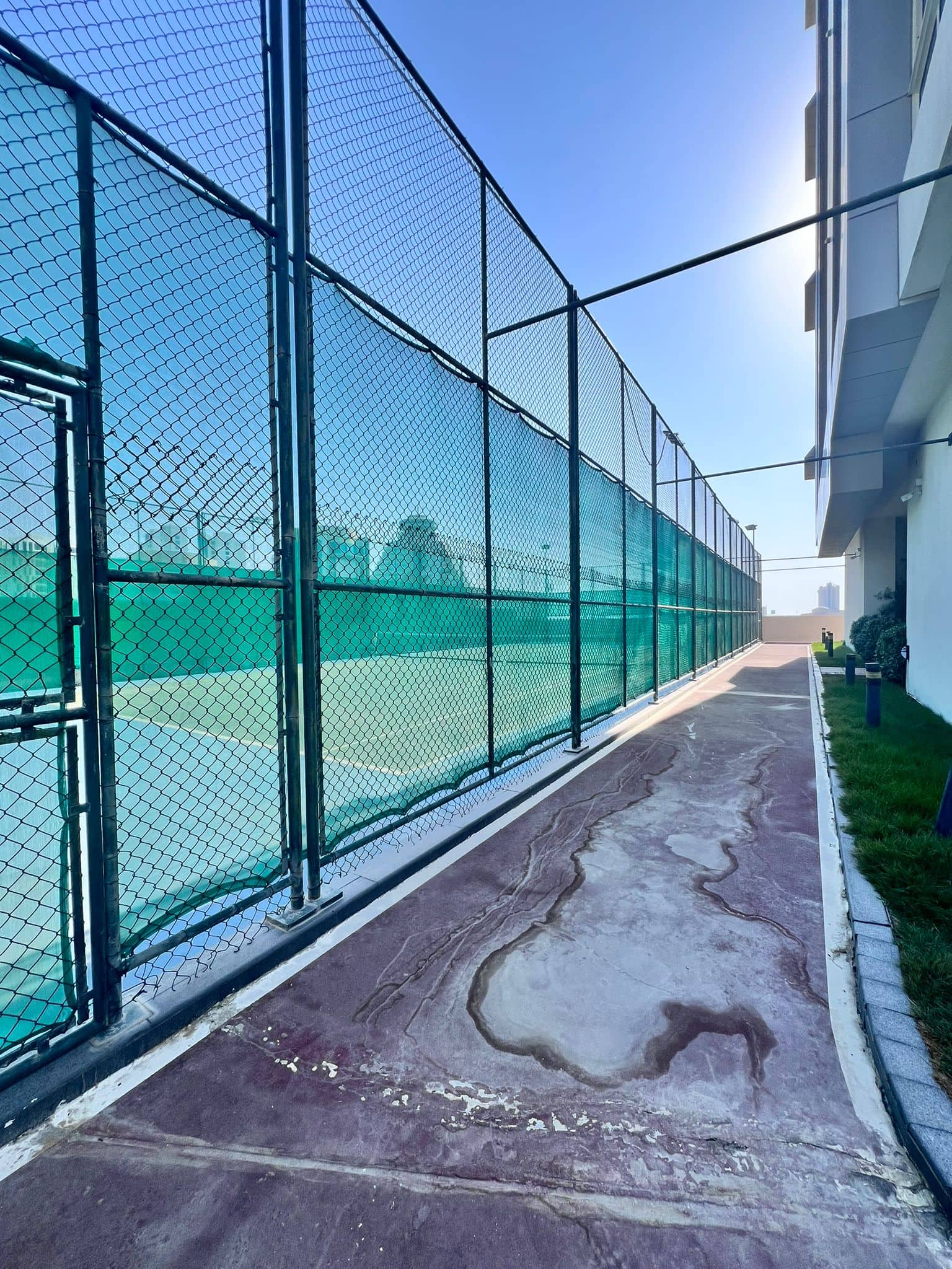 Tennis court with a green fence and weathered path, adjacent to a building under a clear sky.