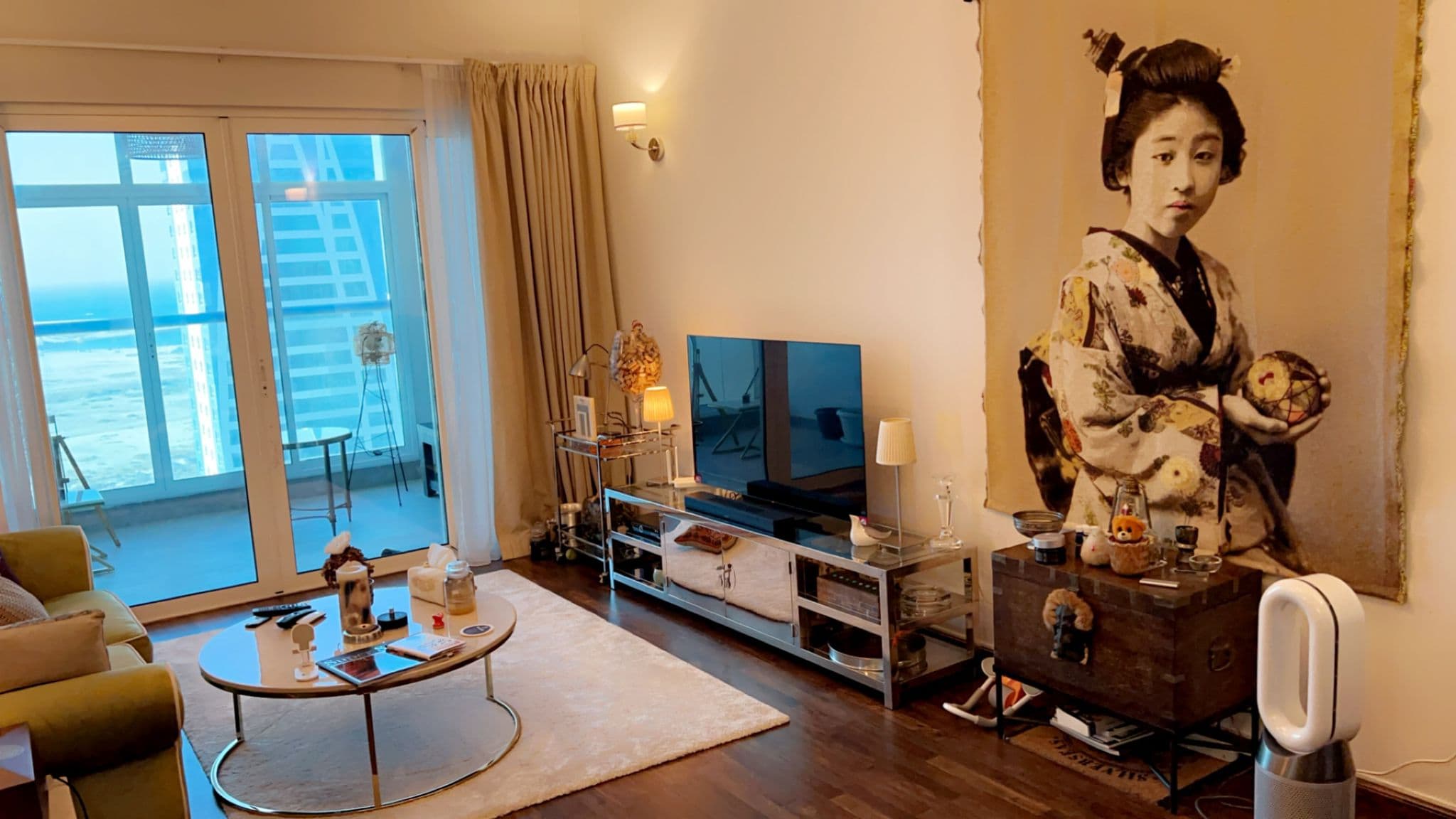 Living room with a large wall art of a woman in traditional attire, a TV on a console, a round coffee table, and floor-to-ceiling windows with a view of high-rise buildings.